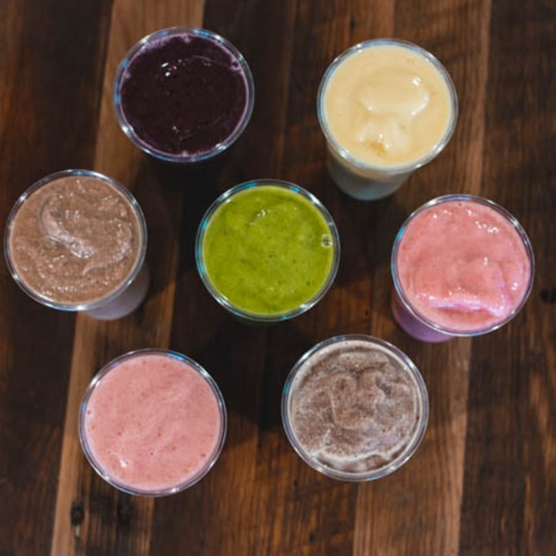 Seven colorful smoothies in clear cups, including purple, yellow, green, pink, and brown, on a wooden table.