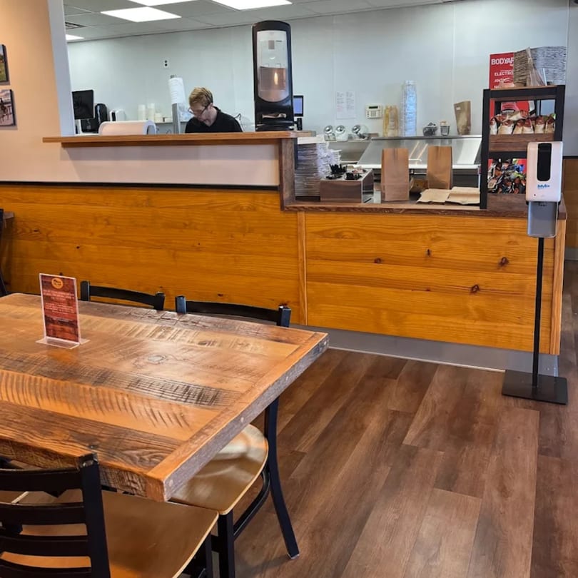 A Mane Course Sandwiches employee works behind a wooden counter, with tables and chairs in the foreground.