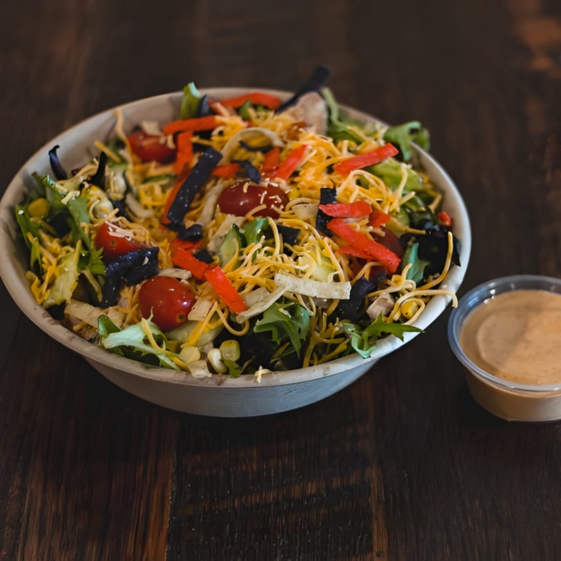 A vibrant salad with mixed greens, cheese, tomatoes, and tortilla strips, with a side of dressing.