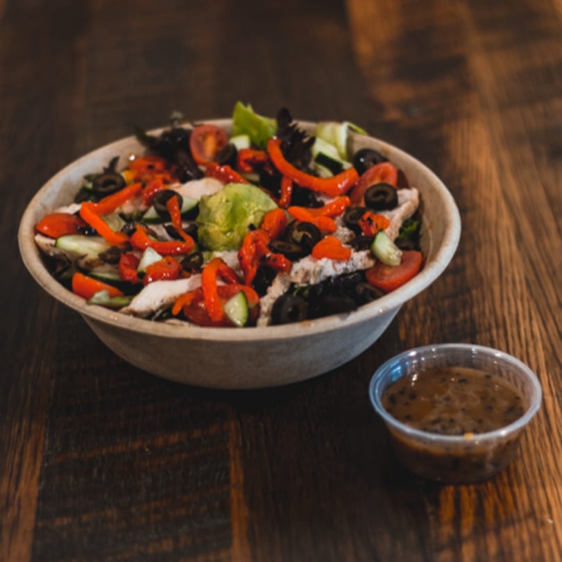 A salad with chicken, red peppers, olives, and greens in a bowl with a side of dressing.