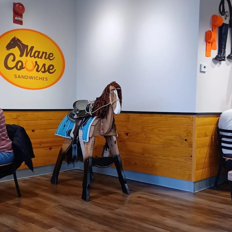 Mane Course Sandwiches restaurant interior with a wooden horse, saddle, and blue blanket, alongside the brand logo.