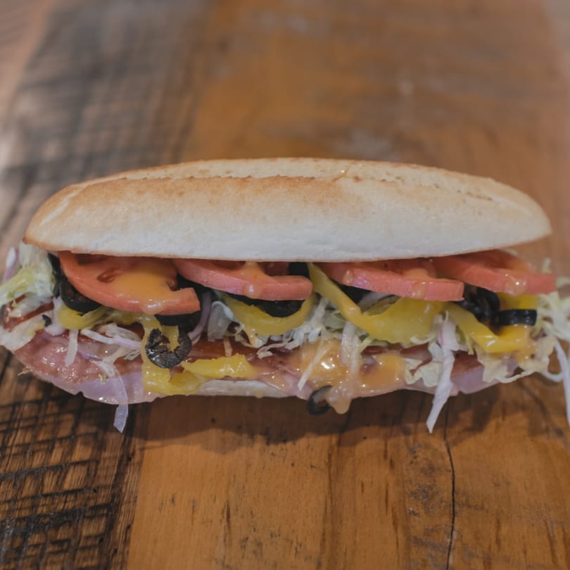 Mane Course Sandwiches' sub sandwich with meat, cheese, tomatoes, olives, and peppers on a wooden table.