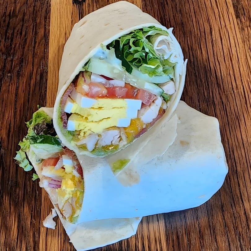 Two halves of a Mane Course sandwich wrap filled with lettuce, tomato, egg, and chicken on a wooden surface.