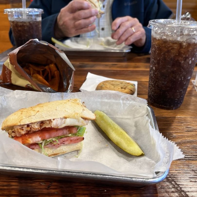 A Mane Course sandwich with a pickle, chips, cookie, and soda on a wooden table.