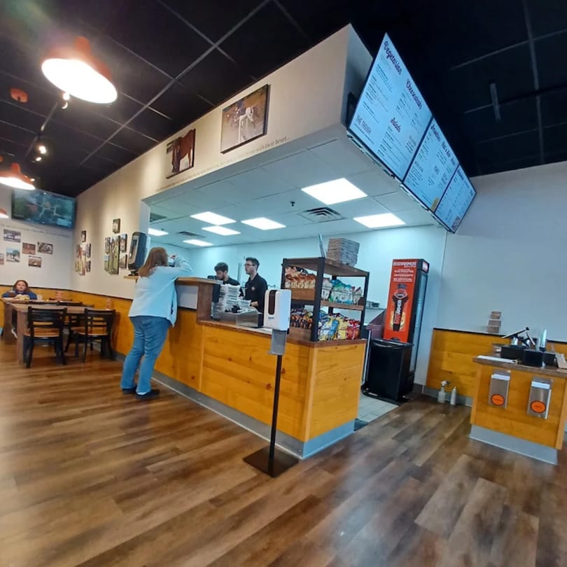 A customer orders at the counter of Mane Course Sandwiches, with staff and menu boards visible.