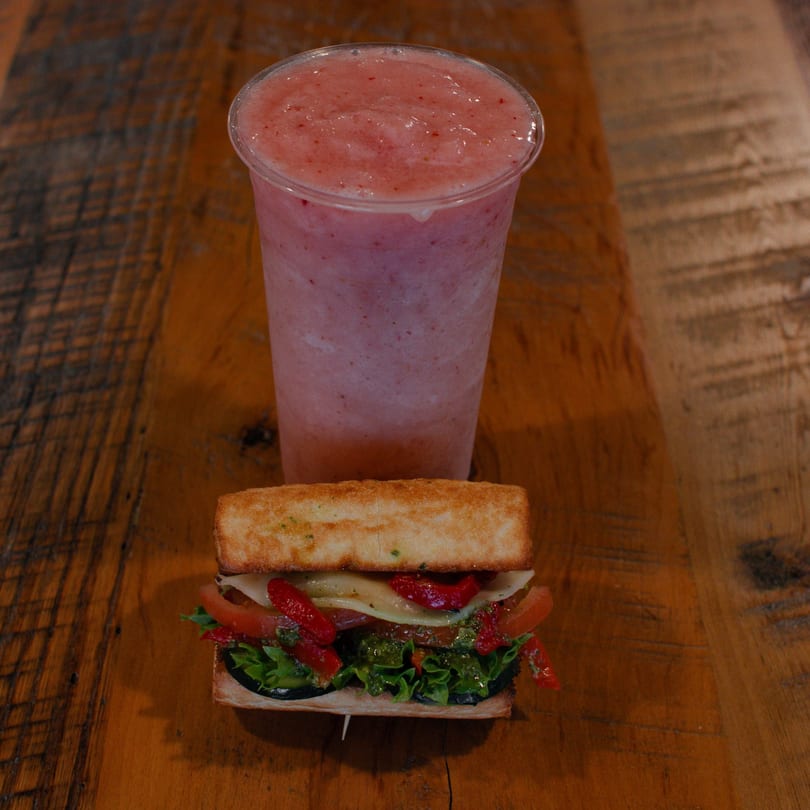 A Mane Course sandwich with lettuce, tomato, cheese, and red peppers, next to a pink smoothie.