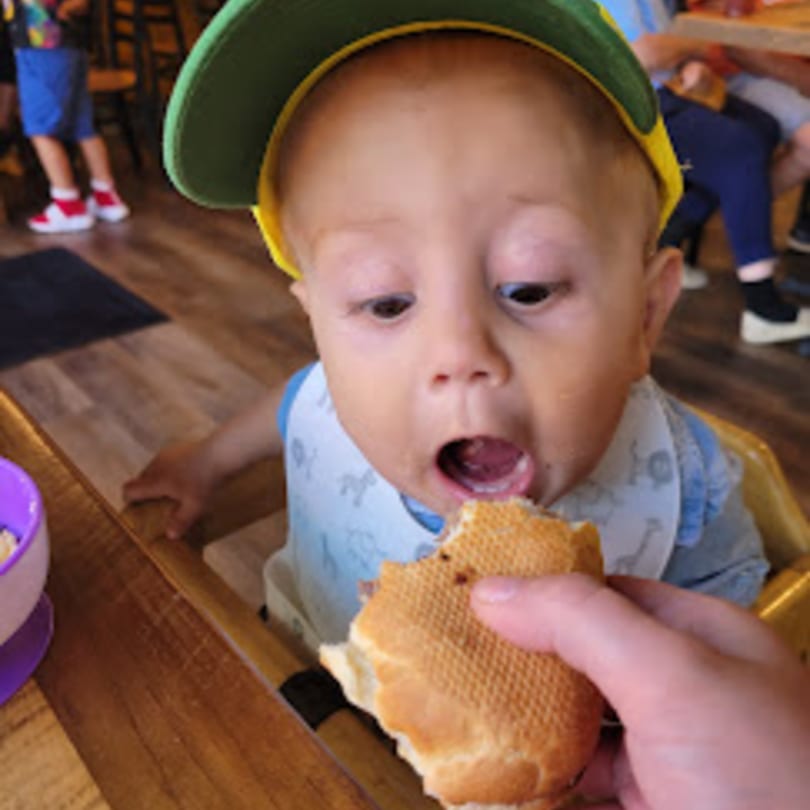 A baby in a high chair with a green hat, mouth open, about to eat a sandwich.