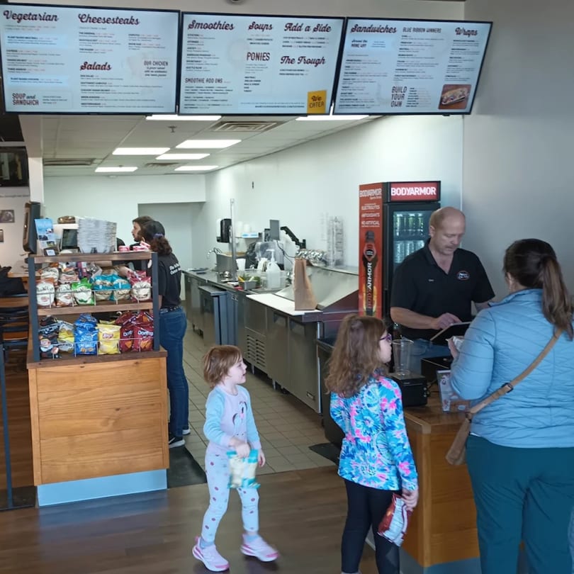 A family with two young girls ordering sandwiches at the counter of Mane Course Sandwiches.